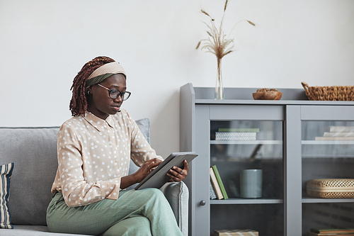 Portrait of young African-American woman using digital tablet while sitting on sofa at home in minimal grey interior, copy space