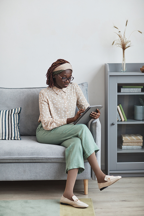 Vertical full length portrait of young African-American woman using digital tablet while sitting on sofa at home in minimal grey interior, copy space