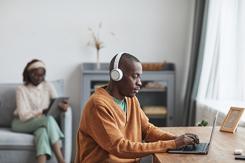 Portrait of contemporary couple using internet devices at home, focus on African-American man wearing headphones at desk in foreground, copy space