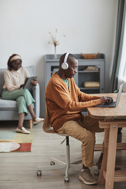 Vertical portrait of contemporary couple using internet devices at home, focus on African-American man wearing headphones and typing at desk in foreground, copy space