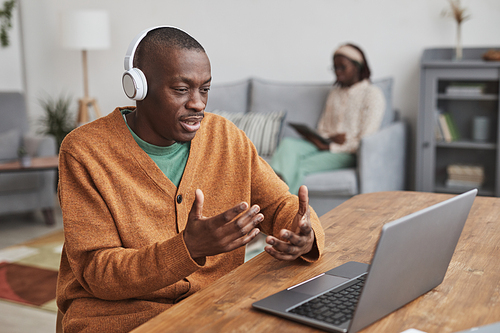 Portrait of African-American man using laptop for video chat and wearing headphones while sitting at desk while in minimal home interior, copy space