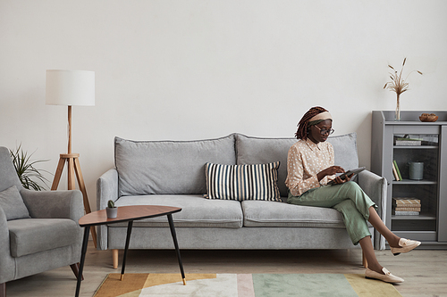 Wide angle portrait of young African-American woman using digital tablet while sitting on sofa at home in minimal grey interior, copy space