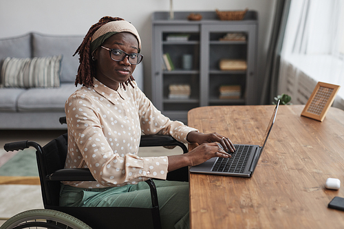 Portrait of young African-American woman using wheelchair and looking at camera while working from home in minimal grey interior, copy space