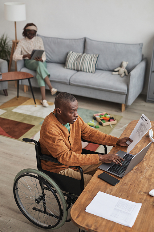 Vertical high angle portrait of African-American man using wheelchair and working from home with childrens toys in background