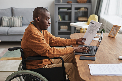 Side view portrait of African-American man using wheelchair working from home in minimal grey interior, copy space