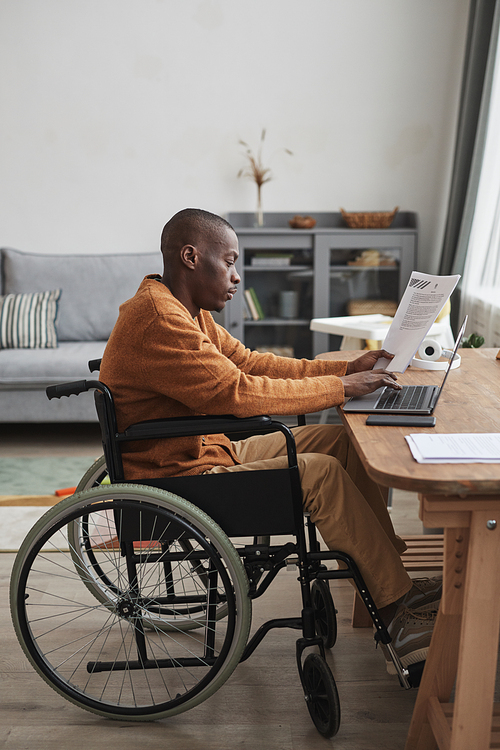 Vertical side view portrait of African-American man using wheelchair working from home in minimal grey interior, copy space