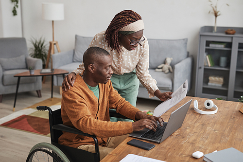 Portrait of African-American man using wheelchair working from home with wife looking over his shoulder, copy space