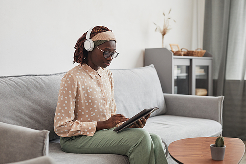 Portrait of young African-American woman wearing headphones and using digital tablet while sitting on sofa at home in minimal grey interior, copy space