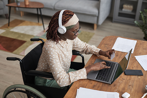 High angle portrait of African-American woman in wheelchair online shopping via laptop and holding credit card, copy space