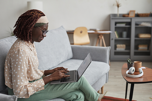 Side view portrait of young African-American woman using laptop while sitting on sofa in modern home interior, copy space