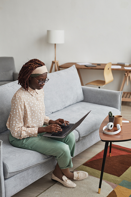 Vertical full length portrait of young African-American woman using laptop while sitting on sofa in minimal home interior, copy space