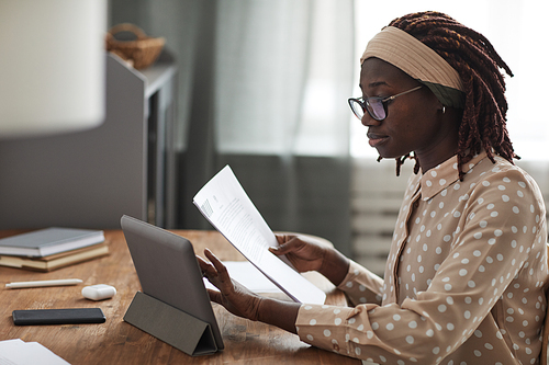 Side view portrait of young African-American woman wearing glasses and using digital tablet on stand while working from home, copy space