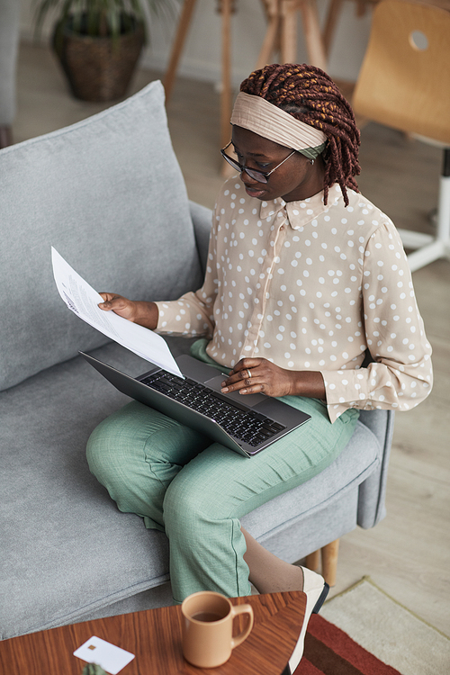 Vertical high angle portrait of young African-American woman wearing glasses and using laptop while sitting on sofa and working from home