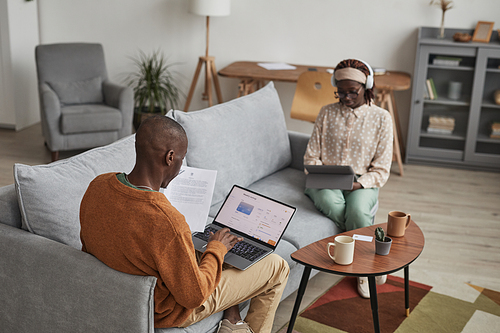 High angle portrait of modern African-American couple using devices while relaxing on sofa at home, focus on man using laptop with online banking service on screen, copy space