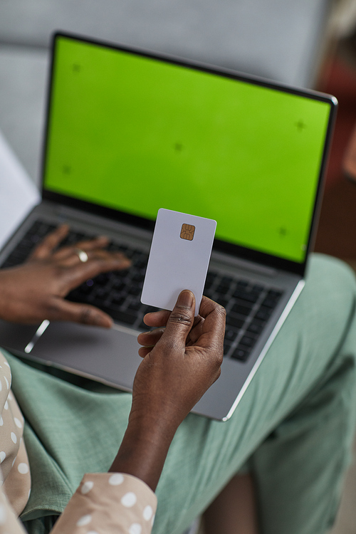 Vertical high angle close up of young African-American woman using laptop with green screen and holding credit card, copy space