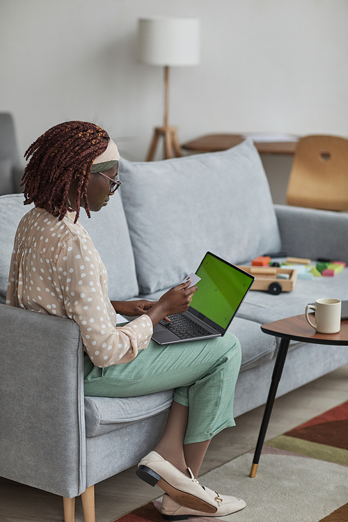 Vertical full length portrait of young African-American woman using laptop with green screen and holding credit card while sitting on sofa at home, copy space