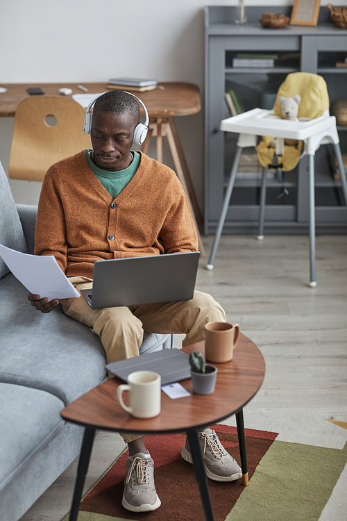 Vertical full length portrait of modern African-American man using laptop and wearing headphones while working from home sitting on sofa