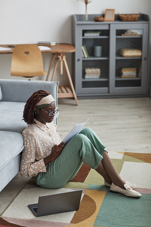Vertical side view portrait of modern African-American woman working from home while sitting on floor at graphic carpet and reading documents, copy space