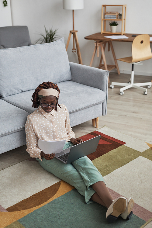 Vertical full length portrait of modern African-American woman working from home while sitting on floor on graphic carpet and using laptop, copy space