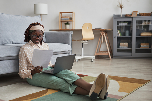 Full length portrait of modern African-American woman working from home while sitting on floor on graphic carpet and using laptop, copy space