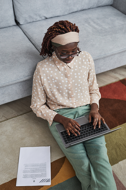 Vertical high angle portrait of modern African-American woman working from home while sitting on floor on graphic carpet and using laptop, copy space