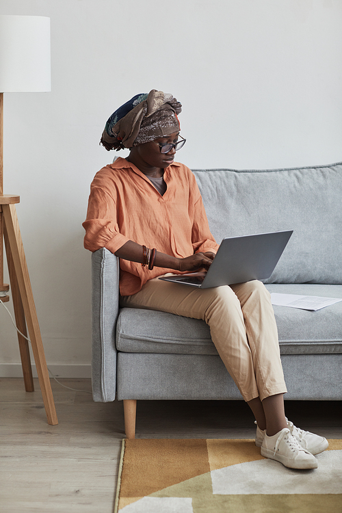 Vertical full length portrait of ethnic Africa-American woman using laptop while sitting on sofa at home, copy space