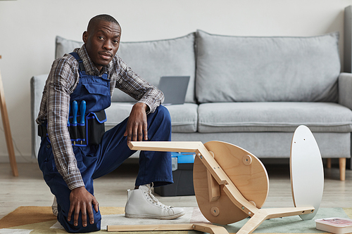 Full length portrait of African-American handyman looking at camera while assembling furniture in home interior, copy space