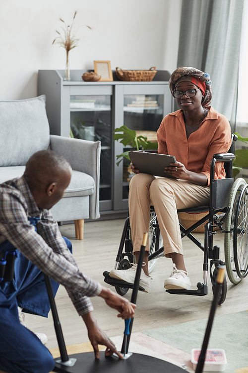 Vertical portrait of young African-American woman in wheelchair watching handyman assembling furniture, online service and assistance concept