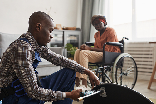 Side view portrait of African-American man assembling furniture in home interior with woman in wheelchair in background, handyman service and assistance concept
