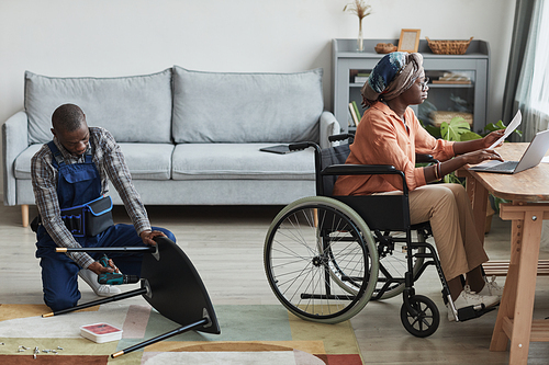 Full length portrait of African-American man assembling furniture in home interior with woman in wheelchair working at desk, handyman service and assistance concept, copy space