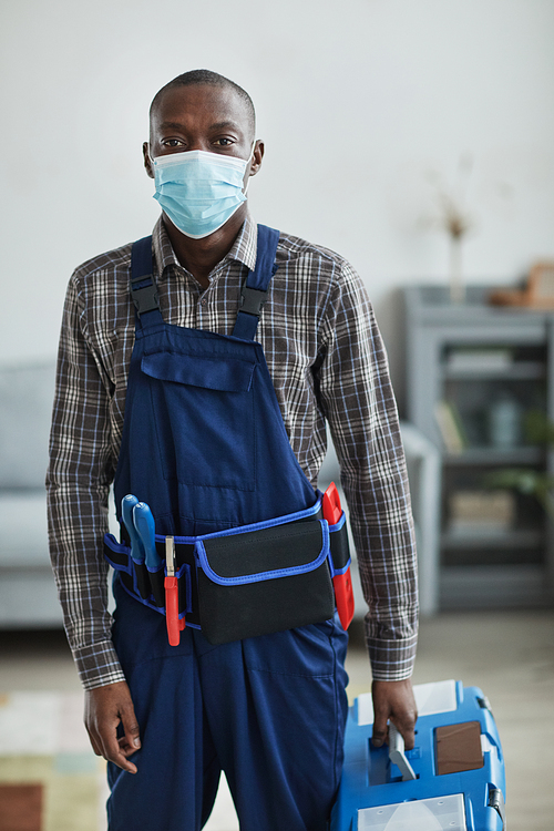 Vertical portrait of African-American handyman wearing mask and holding toolbox while standing in home interior looking at camera