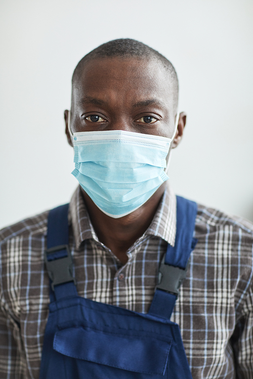 Vertical close up portrait of African-American handyman wearing mask and looking at camera while standing against white wall