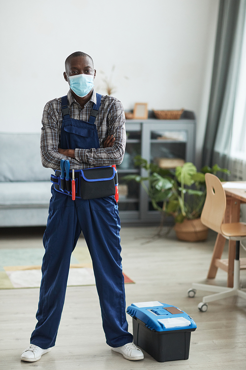 Vertical full length portrait of African-American handyman wearing mask and looking at camera while standing in home interior with toolbox