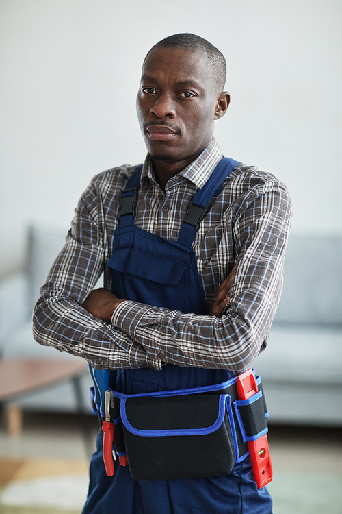Vertical waist up portrait of African-American handyman looking at camera while standing with arms crossed in home interior