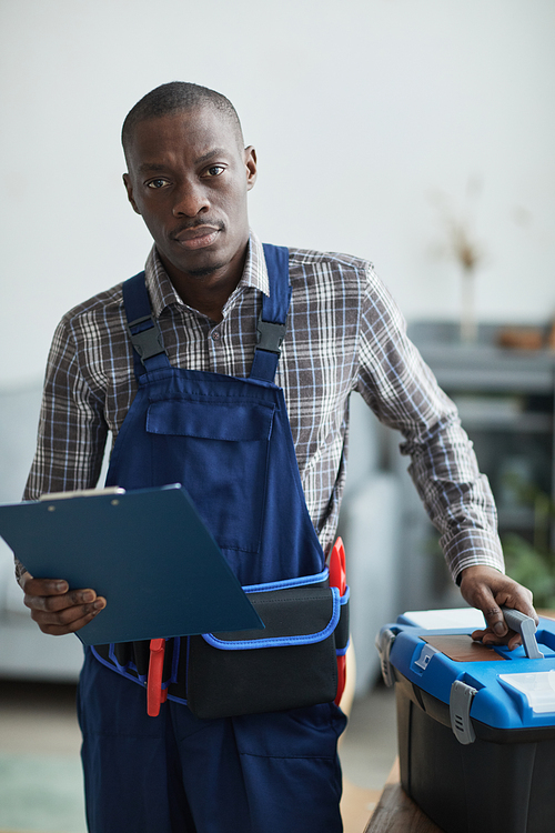 Vertical portrait of African-American handyman looking at camera while standing with toolbox in home interior