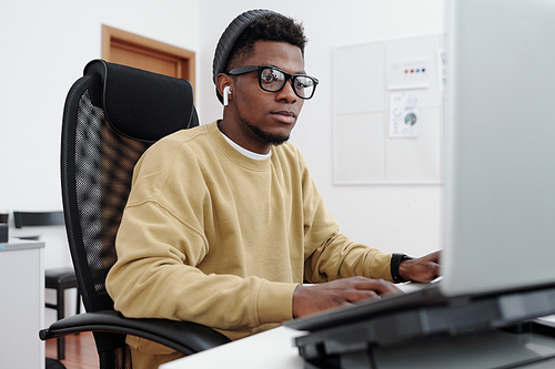 Young contemporary serious male it programmer in yellow sweatshirt looking at computer screen while decoding data in office