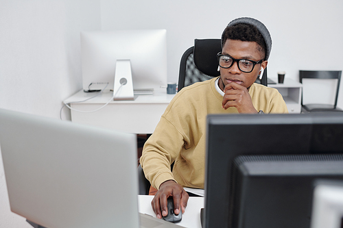 Young serious African American man in casualwear and eyeglasses decoding data in front of computer monitors in office