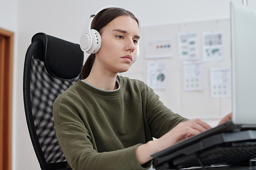 Young serious female programmer in headphones and casualwear decoding data in front of computer monitor by her workplace