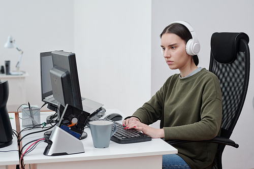 Young serious woman in casualwear listening to music in headphones while sitting by workplace and decoding data in front of computer