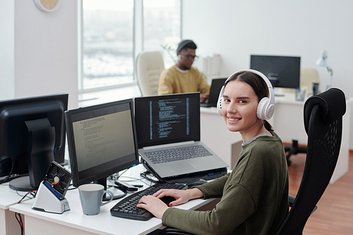 Happy young female it-engineer in headphones sitting by desk in front of computer monitor and laptop and looking at camera in office