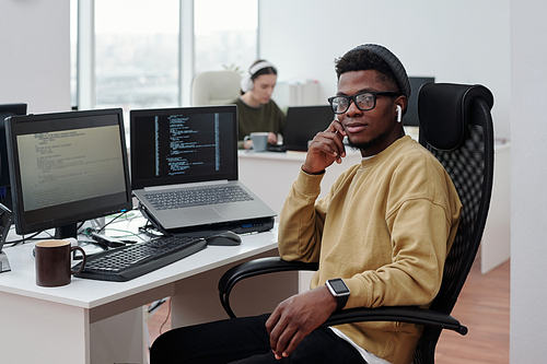 Young confident black man in casualwear with earphones sitting by workplace with compute monitor and laptop with decoded data