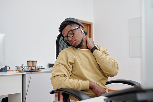Young African American man in casualwear, eyeglasses and earphones doing relaxing exercise for neck by his workplace in office