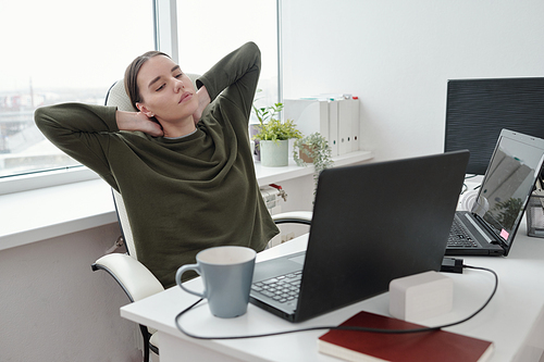 Young relaxed female programmer doing exercise for neck and arms while sitting by her workplace and looking at laptop screen