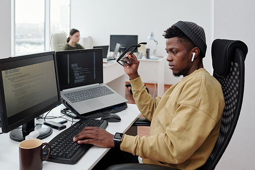 Young contemporary African American it-engineer decoding data by workplace in openspace office while looking at computer screen