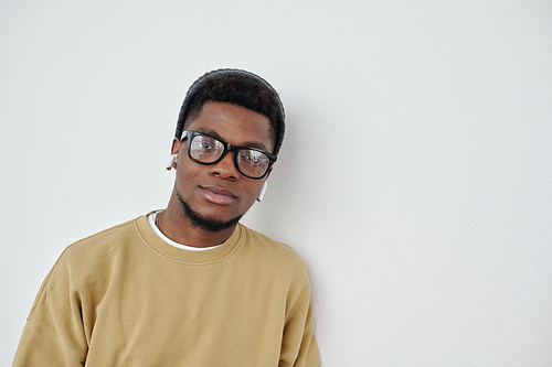 Young confident African American man in eyeglasses, beanie hat and sweatshirt standing by white wall in front of camera