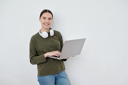 Happy young businesswoman or student with headphones around neck using laptop and looking at camera against white wall in office