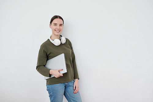 Young contemporary female student with folded laptop standing by white wall and looking at camera with copyspace on her left