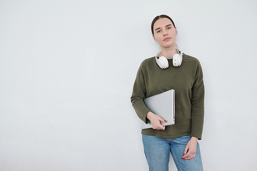 Young serious female student in blue jeans and dark green pullover looking at camera against white wall with copyspace