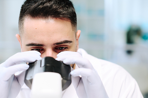Close-up of face of young serious male scientist in gloves looking in microscope during scientific experiment in laboratory