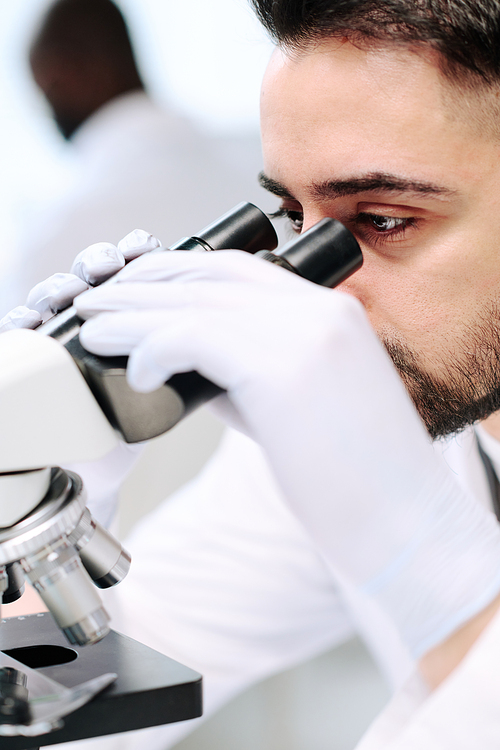 Young male scientist or researcher in gloves studying new virus in microscope while sitting by workplace in modern laboratory